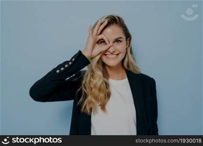 Portrait of beautiful joyful blonde caucasian female wearing black coat looking through okay sign gesture with one eye as if its monocle and smiling, standing alone in front of blue wall. Blonde caucasian female looking through okay sign gesture