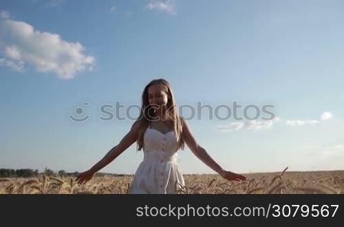 Portrait of beautiful long blond hair woman in white summer dress walking through golden wheat field with cloudy blue sky on background. Attractive smiling female with arms outstretched enjoying freedom and happiness in spikes of wheat field. Slo mo.