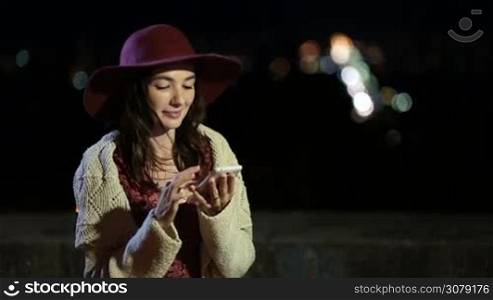 Portrait of beautiful long hair brunette woman in fashionable hat sitting on the bench in park and browsing smartphone at night. Charming millennial female using cell phone with defocused city lights at the background.