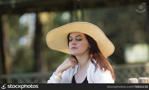 Portrait of beautiful long-haired Hispanic young woman wearing a hat sitting on a park bench with a pensive attitude against a background of unfocused green trees during sunset. Portrait of beautiful long-haired Hispanic young woman wearing a hat sitting on a park bench with a pensive attitude against a background of unfocused green trees