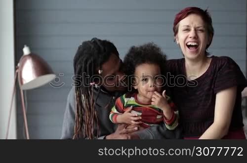 Portrait of beautiful mixed race family looking at camera, smiling and laughing. Positive emotion facial expression. African american father and caucasian mother embracing their adorable mixed race toddler son at home and smiling. Slow motion.