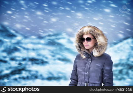 Portrait of beautiful model in winter mountains wearing stylish sunglasses and fashionable coat with fur on the hood enjoying snowfall, wintertime vacation concept