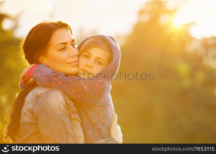 Portrait of beautiful mother with love hugging her precious daughter in bright yellow sunlight in the park, happy family life