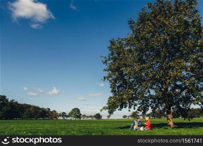 Portrait of beautiful nature and family who has picnic in background sits under big tree. Blue sky and green grass. Charming autumn weather and landscapes. Three people drink tea outdoors on meadow
