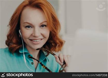 Portrait of beautiful redhead professional female teaches conducts lessons online, waits for starting video conference with pupils, uses earphones and notebook, smiles positively, holds spectacles