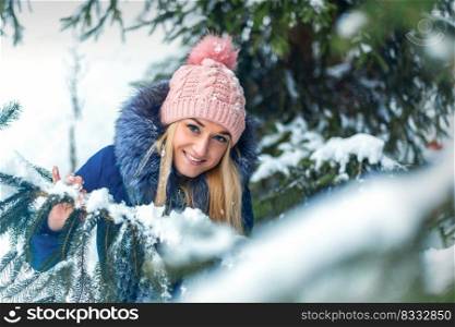 Portrait of beautiful smiling caucasian young woman standing in winter forest wearing blue jacket and pink hat. Young woman standing in winter forest