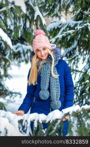 Portrait of beautiful smiling caucasian young woman standing in winter forest wearing blue jacket and pink hat. Young woman standing in winter forest