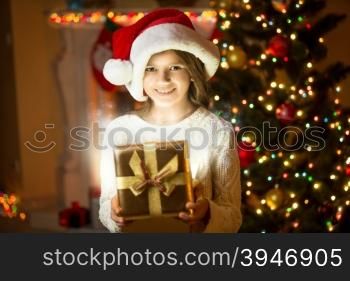 Portrait of beautiful smiling girl posing with shining gift box against Christmas tree