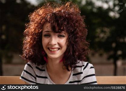 Portrait of beautiful smiling girl with curly red hair close up, against background of summer city park.