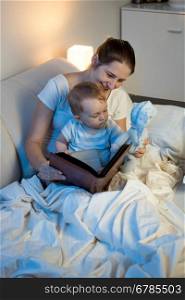 Portrait of beautiful smiling mother reading fairytale book to her baby at bed