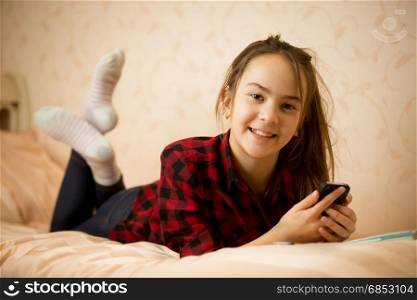 Portrait of beautiful smiling teenage girl ling on bed and using smartphone