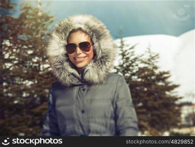 Portrait of beautiful smiling woman in the snowy mountains, wearing warm coat with fur hood and stylish sunglasses, happy winter holidays