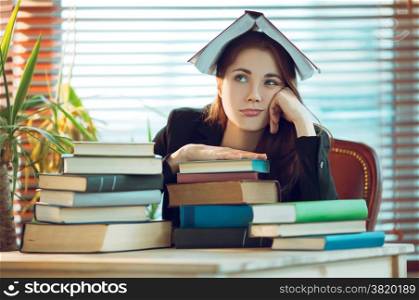 Portrait of beautiful student girl sitting among books with a book on her head