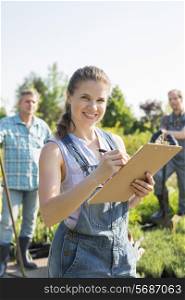 Portrait of beautiful supervisor holding clipboard with gardeners standing in background at plant nursery