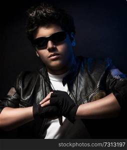 Portrait of beautiful teen boy wearing stylish sunglasses, leather jacket and special sportive gloves over blue background, fashion accessories for teenagers