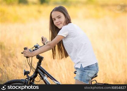 Portrait of beautiful teenage girl posing on bicycle at meadow at sunset