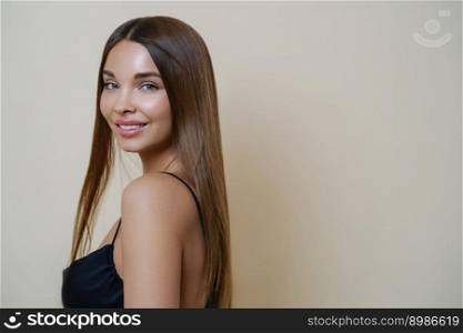 Portrait of beautiful tender girl with well cared long dark hair, has bare shoulder, dressed in black dress, has pleased expression, isolated over beige background. Beauty, makeup, personal care