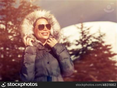 Portrait of beautiful woman having fun in the winter park, wearing sunglasses and coat with fur hood in frosty sunny day, stylish wintertime look