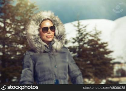Portrait of beautiful woman in the winter mountains, wearing warm coat with fur hood and stylish sunglasses