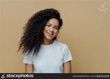 Portrait of beautiful woman with frizzy bushy hair, gentle smile and healthy skin, wears casual white t shirt, enjoys pleasant conversation stands against beige background copy space. People, emotions