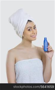 Portrait of beautiful woman wrapped in towel holding hair oil bottle against gray background