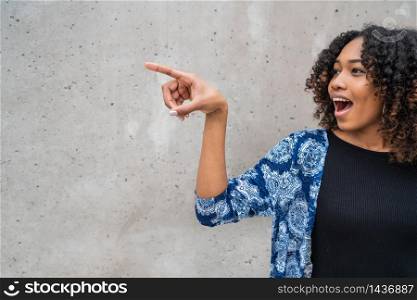Portrait of beautiful young afro-american woman with curly hair pointing something against grey wall.