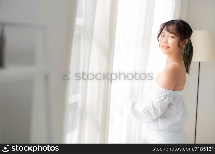 Portrait of beautiful young asian woman standing the window and smile while wake up with sunrise at morning, girl happy with freshness and cheerful, lifestyle and relax concept.