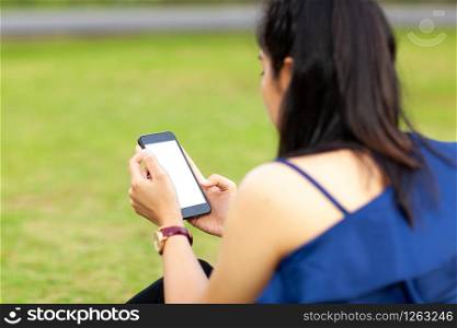 Portrait of beautiful young asian woman using blank display of smartphone and sitting on green grass field in the public park included clipping path