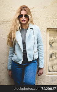 Portrait of beautiful young blonde woman with curly hair. Girl wearing leather jacket, blue jeans and aviator sunglasses in urban background.