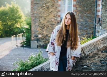 Portrait of beautiful young female model wears warm mantlet, keeps hands in pocket, poses against ancient landmarks, enjoys wonderful nature and calm atmosphere, admires archtectural builduings