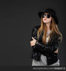 Portrait of beautiful young girl wearing black felt hat, Sunglasses and leather jacket in front of a dark gray background