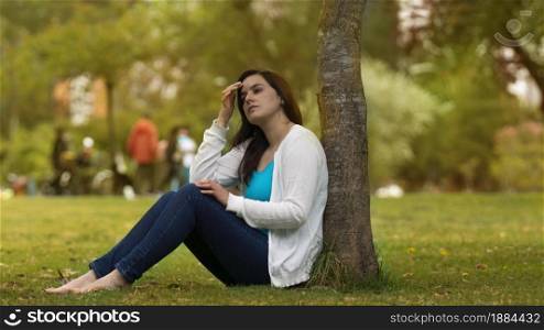 Portrait of beautiful young Hispanic woman sitting under a tree in the middle of a park with a pensive attitude against a background of unfocused trees during the day. Portrait of beautiful young Hispanic woman sitting under a tree in the middle of a park with a pensive attitude against a background of unfocused trees