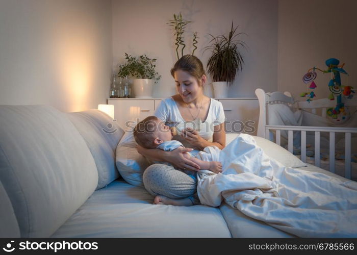 Portrait of beautiful young mother giving milk to her baby boy in bed at night