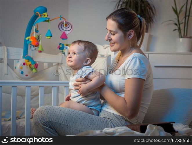 Portrait of beautiful young mother sitting on bed at night and holding adorable baby boy