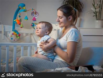 Portrait of beautiful young mother sitting on bed at night and holding adorable baby boy