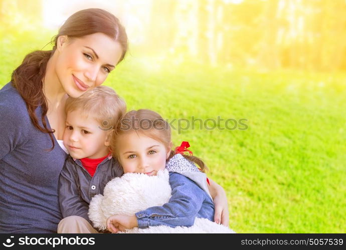 Portrait of beautiful young mother with cute daughter and son sitting on the meadow, sunny day, love and happy family concept