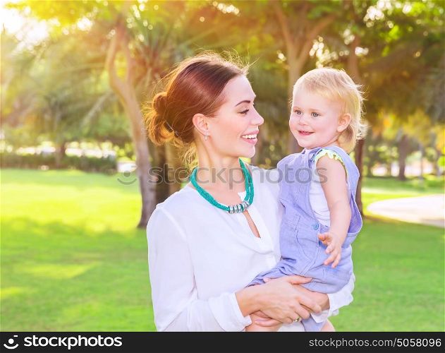 Portrait of beautiful young mother with cute little daughter on hands spending summer day in fresh green park, having fun outdoors, love and happiness concept