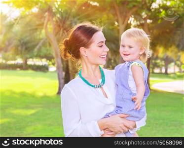 Portrait of beautiful young mother with cute little daughter on hands spending summer day in fresh green park, having fun outdoors, love and happiness concept