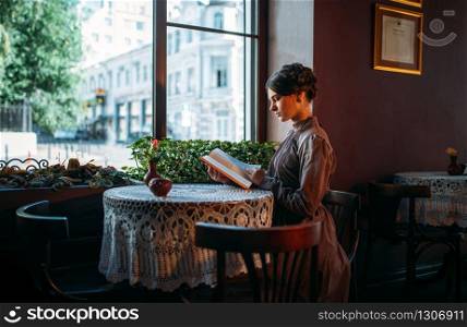 Portrait of beautiful young retro lady in cafe reading book near window. Adorable young lady having rest in vintage cafe with book. Serious brunette in dress dark gray color.. Portrait of lady in cafe reading book near window