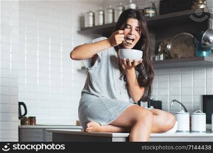 Portrait of beautiful young woman eating bowl of cereal and fruit at home in kitchen.