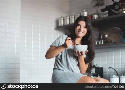 Portrait of beautiful young woman eating bowl of cereal and fruit at home in kitchen.