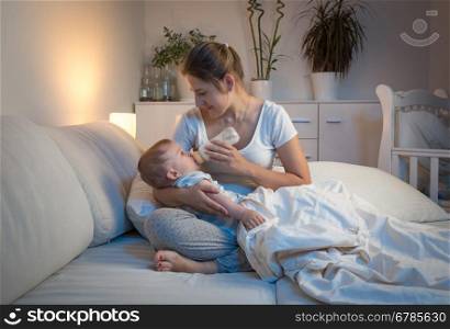 Portrait of beautiful young woman giving milk from bottle to her baby at night
