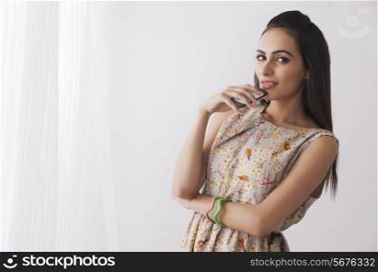 Portrait of beautiful young woman holding cell phone by curtains