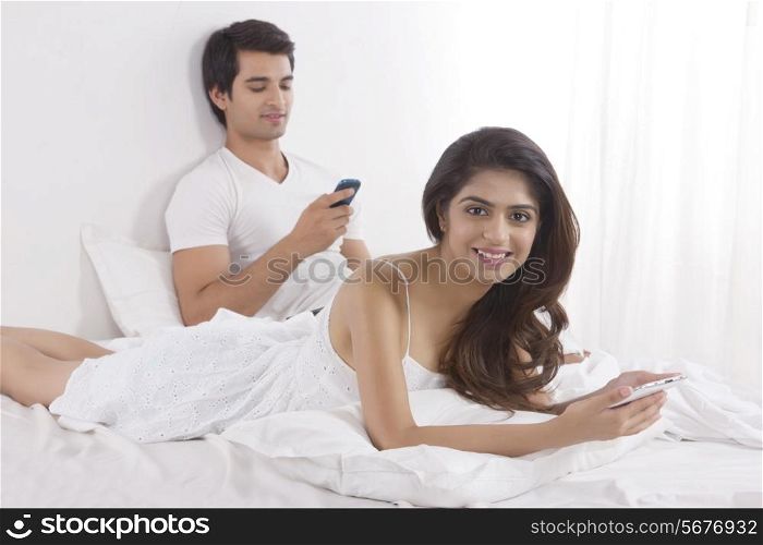 Portrait of beautiful young woman holding digital tablet with man using cell phone in bed
