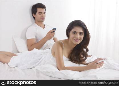 Portrait of beautiful young woman holding digital tablet with man using cell phone in bed