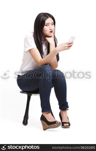 Portrait of beautiful young woman in casuals sitting on stool with mobile phone against white background