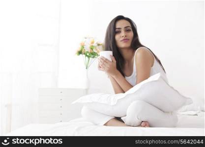 Portrait of beautiful young woman in nightwear holding coffee cup on bed