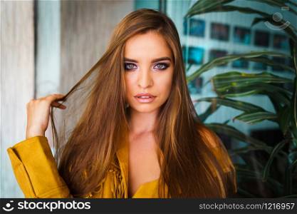 Portrait of beautiful young woman model brunette holding long hair posing by the window in yellow jacket