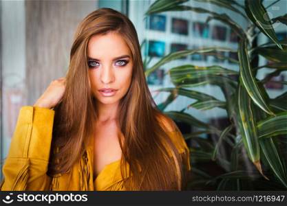 Portrait of beautiful young woman model brunette with long hair posing by the window in yellow jacket