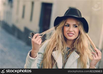 Portrait of beautiful young woman playing with her curly hair and smiling in urban background. Girl wearing jacket and hat.